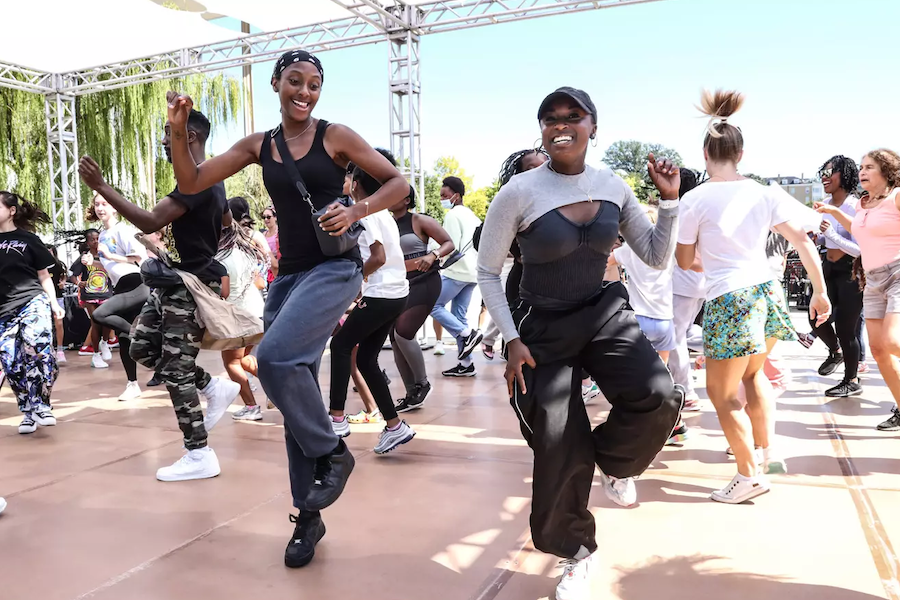  A group of people energetically dancing together outdoors under a canopy, led by two women in the foreground who are smiling and having fun.