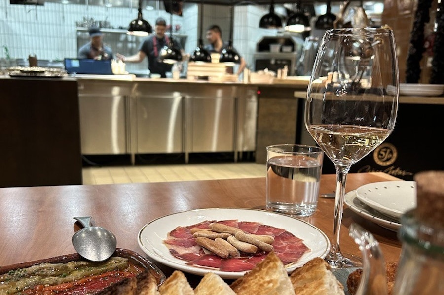 a dining table with plates of Italian-style food and a wine glass overlooking a restaurant chef's station