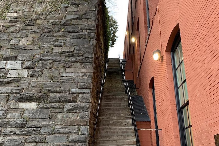 A steep staircase between a stone wall and a red brick building, with lights mounted on the building illuminating the steps.