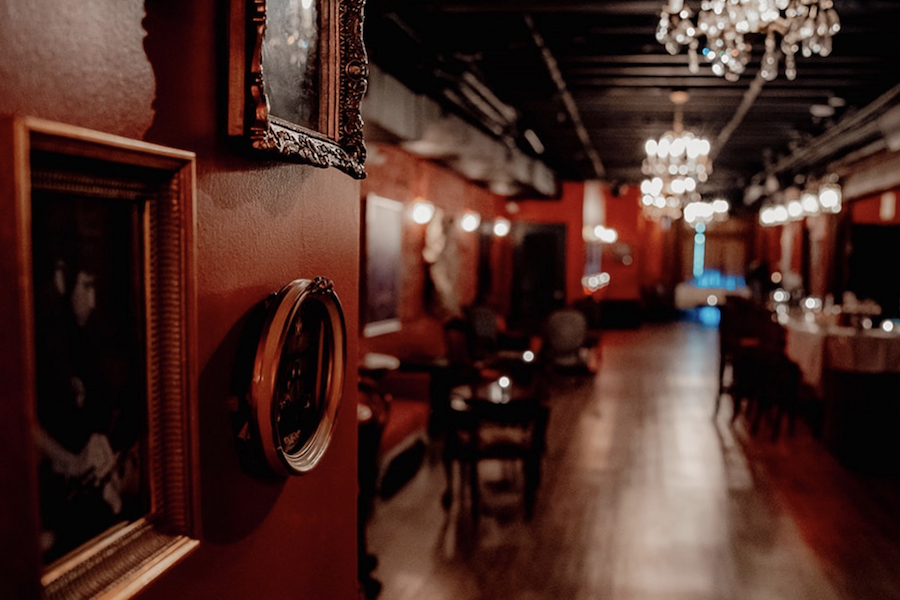 A dimly lit, elegant hallway with chandeliers hanging from the ceiling, framed pictures on the red walls, and antique furniture placed along the sides.