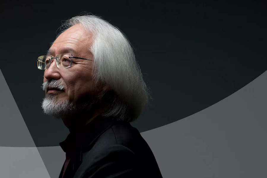 A side profile of a man with white hair and a beard, wearing glasses and a black suit, set against a dark, minimalistic background.