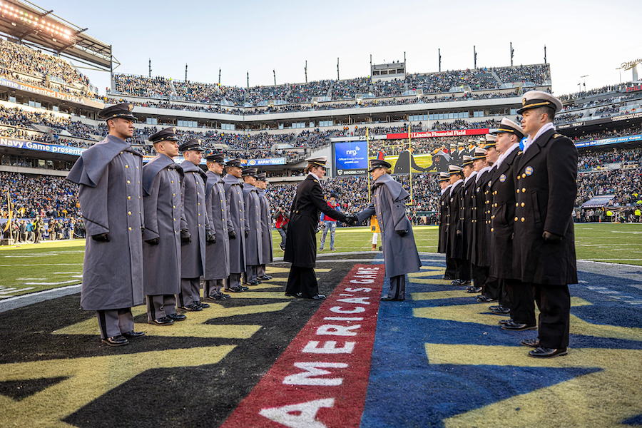 Cadets from Army and Navy stand in formation facing each other on the football field during the Army-Navy game, with two representatives shaking hands at midfield. The stadium is filled with spectators, and the words 'America's Game' are visible on the field.