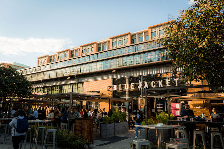  Exterior view of Bluejacket brewery in Washington, DC, with people enjoying drinks at outdoor tables under a clear sky.