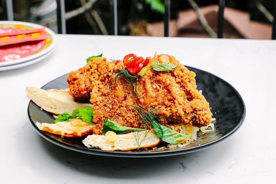 A crispy fried chicken dish garnished with herbs and sliced peppers, served with bread on a black plate.