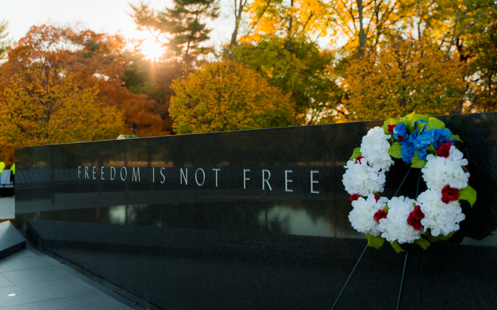 A solemn scene at a memorial, featuring a stone wall engraved with the words "FREEDOM IS NOT FREE." A wreath of red, white, and blue flowers rests against the wall, with autumn trees and the warm glow of a setting sun in the background.