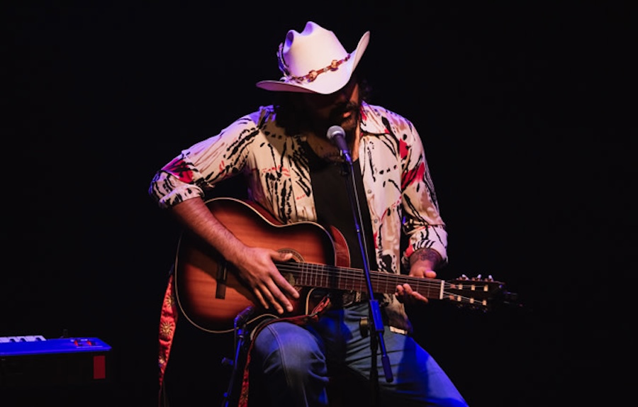  A musician wearing a cowboy hat and patterned shirt performs on stage, playing an acoustic guitar and singing into a microphone under a spotlight.