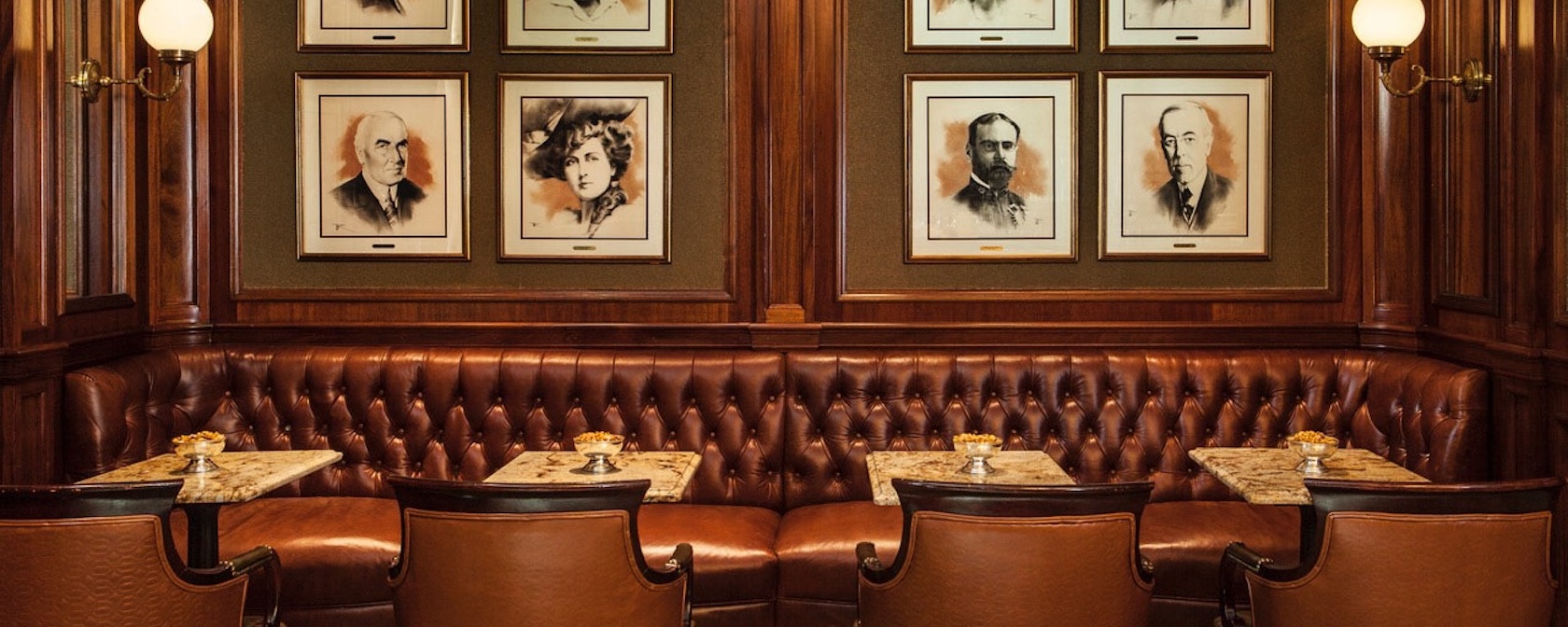 A cozy seating area featuring tufted leather banquettes, small marble tables, and portraits of notable historical figures adorning the walls in a warm wooden setting.