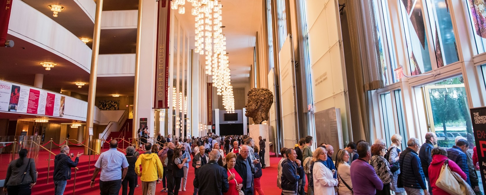 A crowded lobby inside the Kennedy Center, with people walking past a large sculpture and chandeliers under a high ceiling.