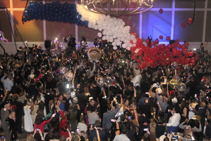 A festive ballroom scene filled with a lively crowd celebrating under a cascade of red, white, and blue balloons. Guests are dressed elegantly, many holding phones to capture the moment as lights and decorations create a vibrant atmosphere.