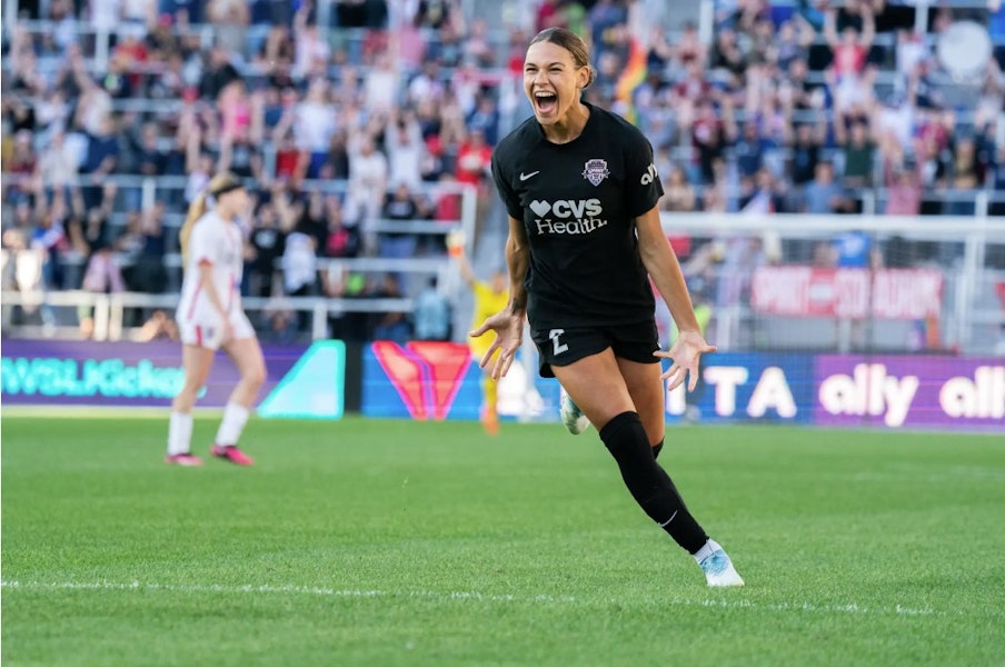 A Washington Spirit player celebrates a goal with an intense expression as the crowd cheers in the background.