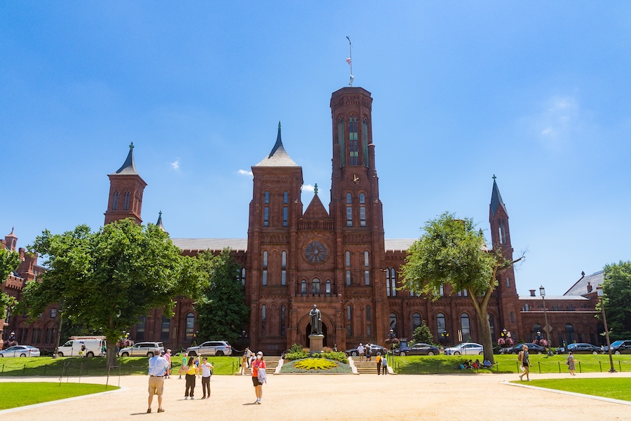 Visitors walk across the National Mall toward the red sandstone Smithsonian Castle on a sunny day.