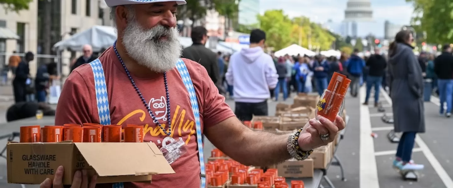 A man with a white beard and a blue checkered hat and suspenders, handing out orange beer cans at an Oktoberfest event in Washington D.C., with the U.S. Capitol building visible in the background.