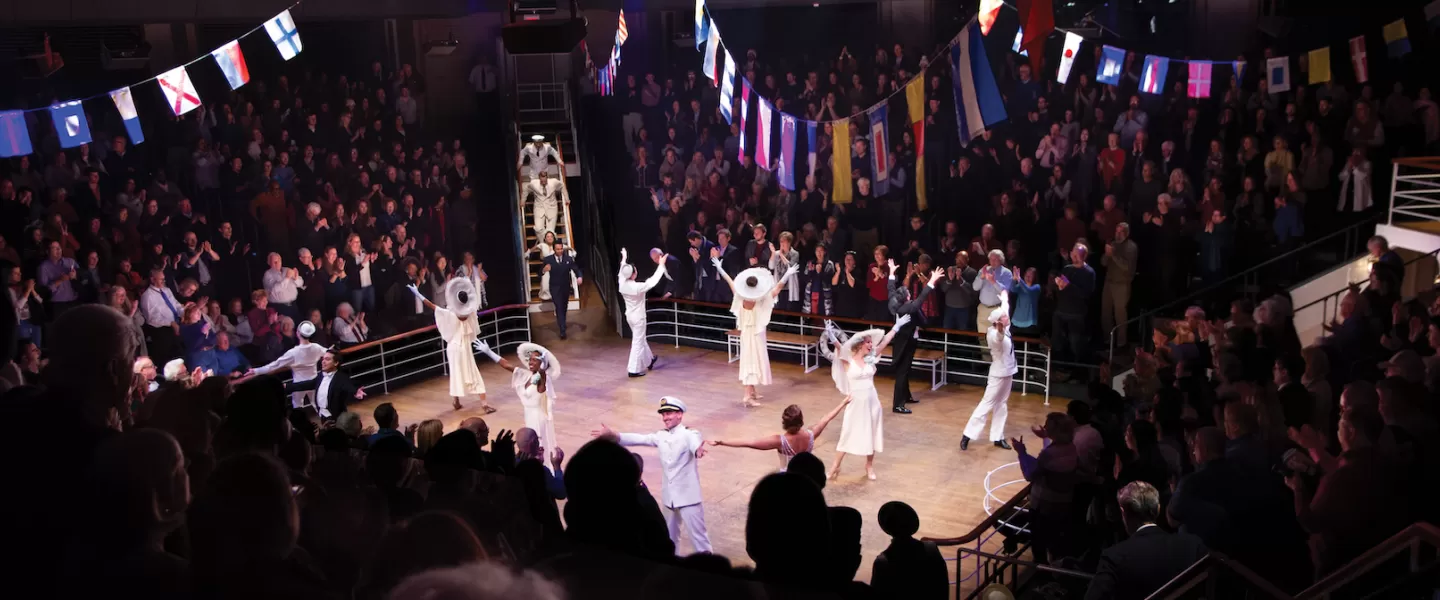 A lively scene from "Anything Goes" with actors in 1930s-style costumes on a ship deck set. The audience claps enthusiastically, surrounded by colorful flags hanging overhead.