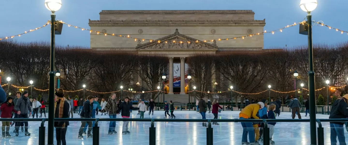 People ice skating at the National Gallery of Art Sculpture Garden rink during the evening, with string lights overhead and the museum building in the background.