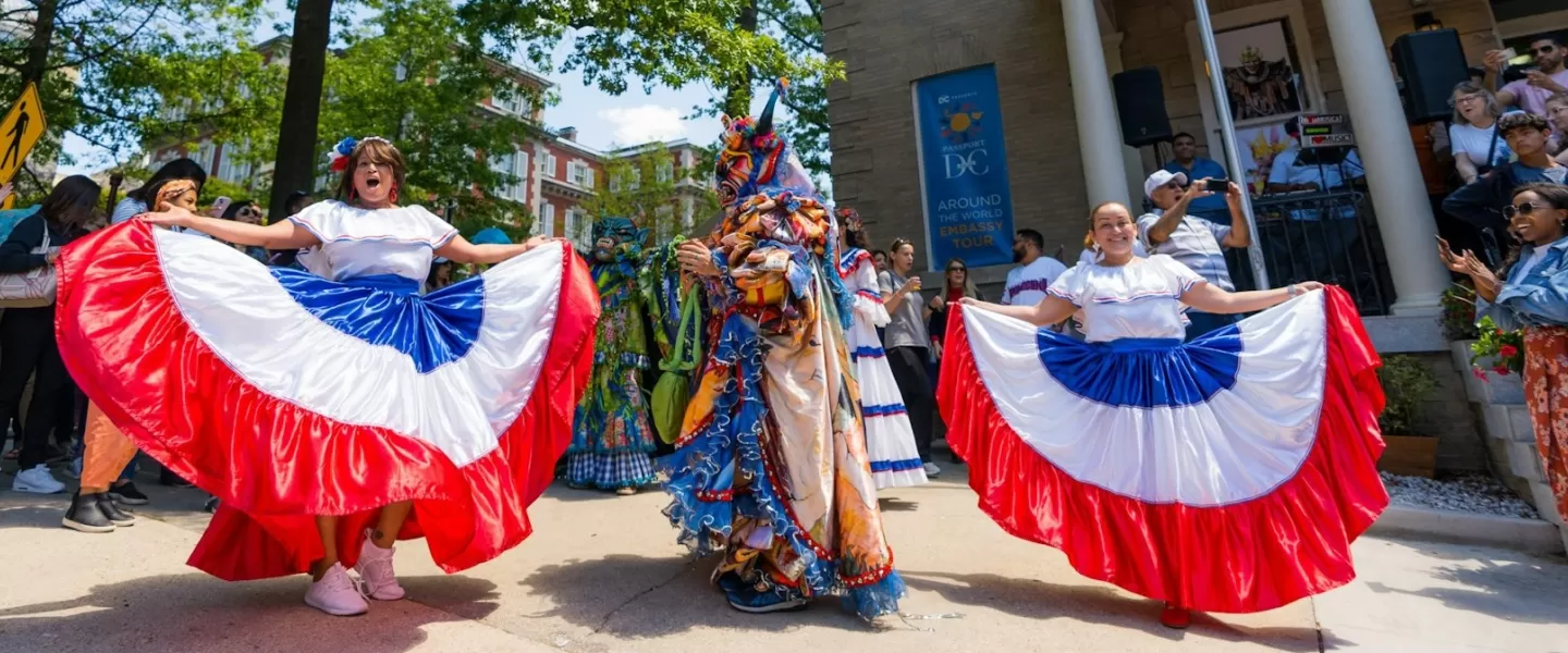 Two women in colorful skirts dance alongside a costumed figure, celebrating embassies in Washington, DC. 