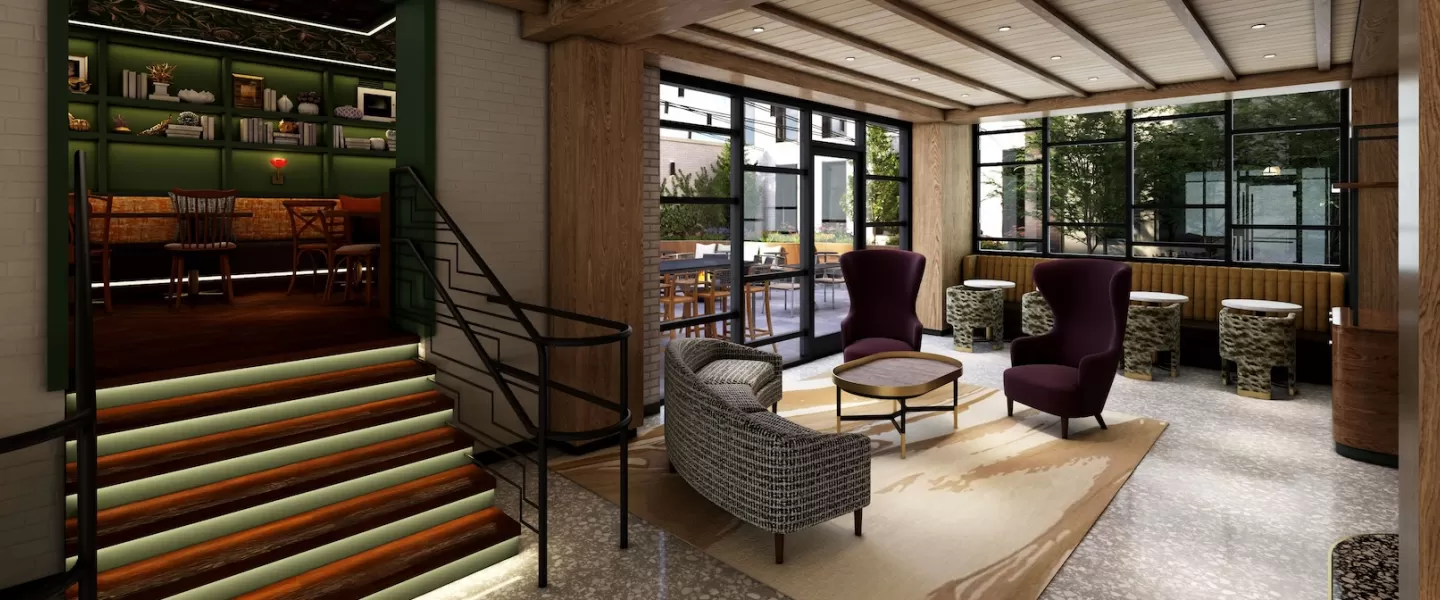 A stylish hotel lobby featuring a mix of modern and classic furnishings, with cozy lounge chairs, a carpeted seating area, a staircase leading to a book-lined nook and large windows letting in natural light.
