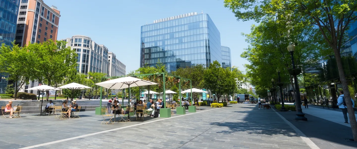 A lively outdoor plaza with people sitting under white umbrellas surrounded by modern glass buildings and leafy green trees on a sunny day.