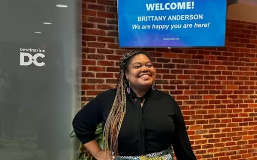 Brittany Anderson, a Black woman with long braids, stands with a smile in front of a sign that says WELCOME!

