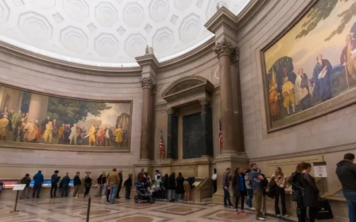Visitors line up inside the National Archives Rotunda beneath historic murals and a domed ceiling.
