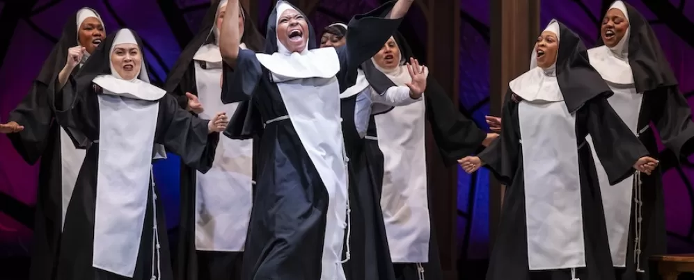 A nun holding a conductor ’ s wand dances animatedly with arms stretched up in the air as a large group of nuns dance and sing behind her.