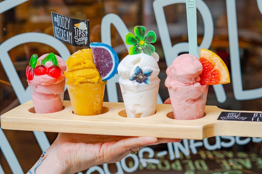 A hand holding a wooden tray with four small cups of colorful, decorated boozy ice cream scoops from Tipsy Scoop. Each scoop is topped with garnishes like cherries, citrus slices, and candy decorations, displayed against a window with part of the "Boozy Ice Cream" sign visible in the background.