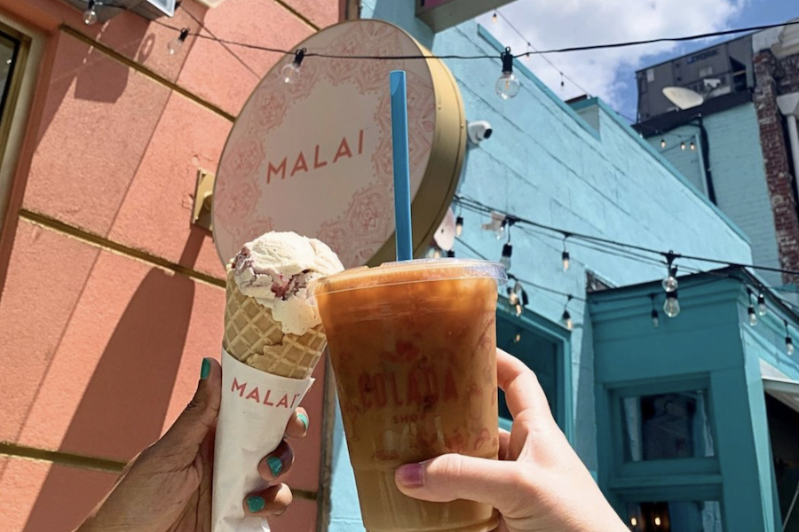 A hand holding an ice cream cone and another hand holding an iced coffee in front of Malai ice cream shop, with a colorful wall and sign visible in the background.