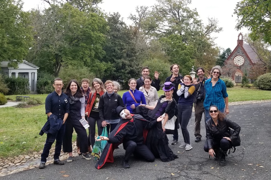 A group of people posing outdoors in a park-like setting, with two individuals dressed in theatrical costumes, including a vampire cape and a purple top hat. The group is smiling and laughing, with trees and a small chapel in the background.