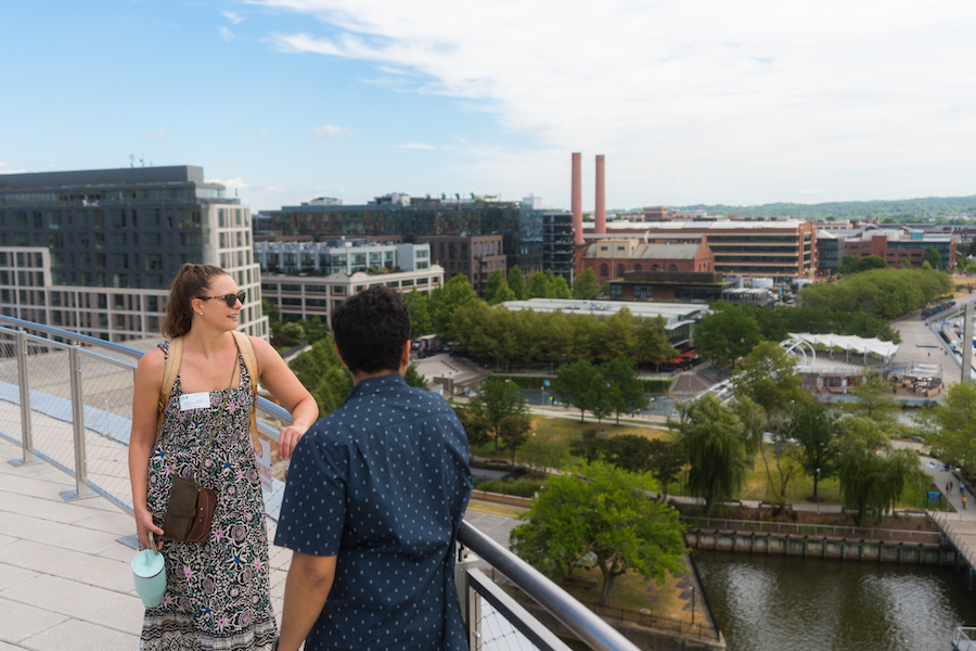 A woman in a patterned dress and sunglasses talks to a man in a blue shirt on a rooftop terrace, overlooking a cityscape with modern buildings and greenery under a partly cloudy sky.