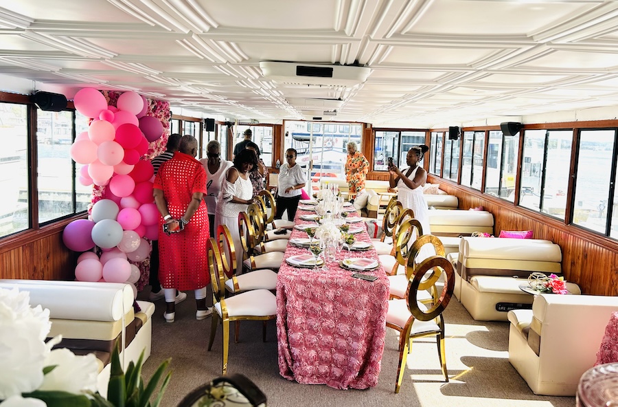 A beautifully decorated dining setup with pink accents and balloon arrangements on a boat, as guests in elegant attire prepare for the event.