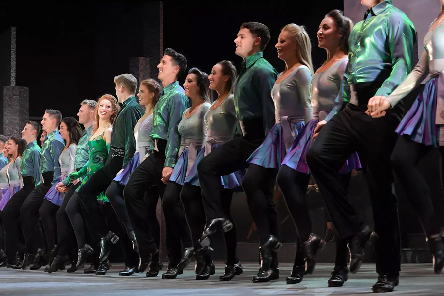 A line of dancers in vibrant costumes perform a synchronized Riverdance routine on stage.