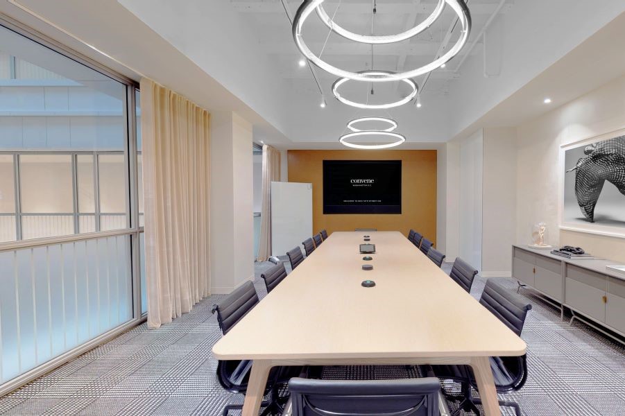 A sleek boardroom with a long wooden table, black chairs, circular overhead lights, a wall-mounted screen, and modern decor, creating a professional meeting environment.