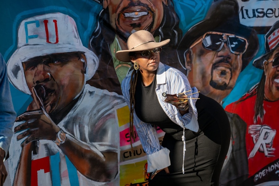 A woman in sunglasses and a hat poses in front of a colorful mural celebrating go-go musicians in Washington, DC.