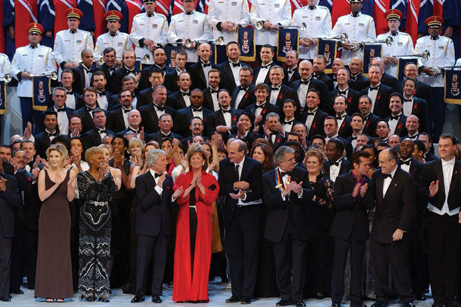 Large group of performers and dignitaries in formal attire on stage, applauding during a ceremony.