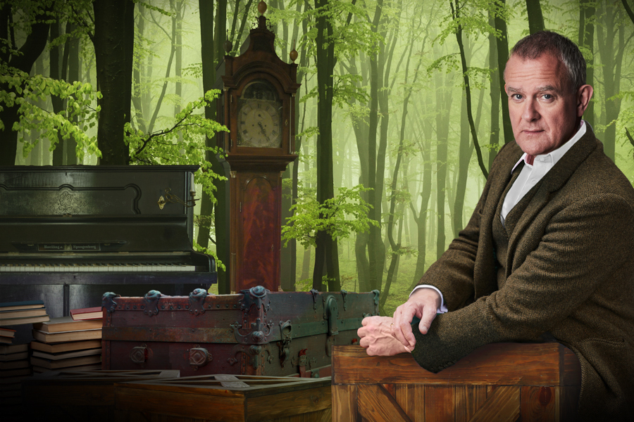 Man in a tweed jacket poses among vintage items, including a piano, clock, books, and trunks, set against a lush green forest backdrop.