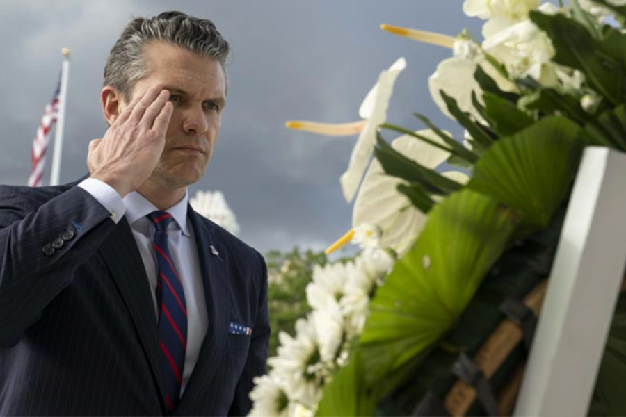A man in a suit salutes a ceremonial wreath in front of an American flag during a solemn Memorial Day event.