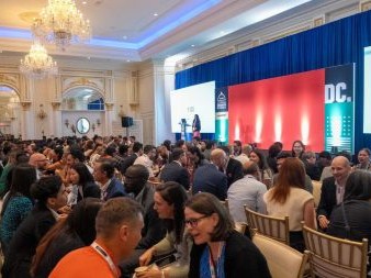 A lively networking session in a grand ballroom with chandeliers, where rows of attendees sit face-to-face engaging in timed conversations while a speaker stands on stage in front of a large red and blue backdrop.