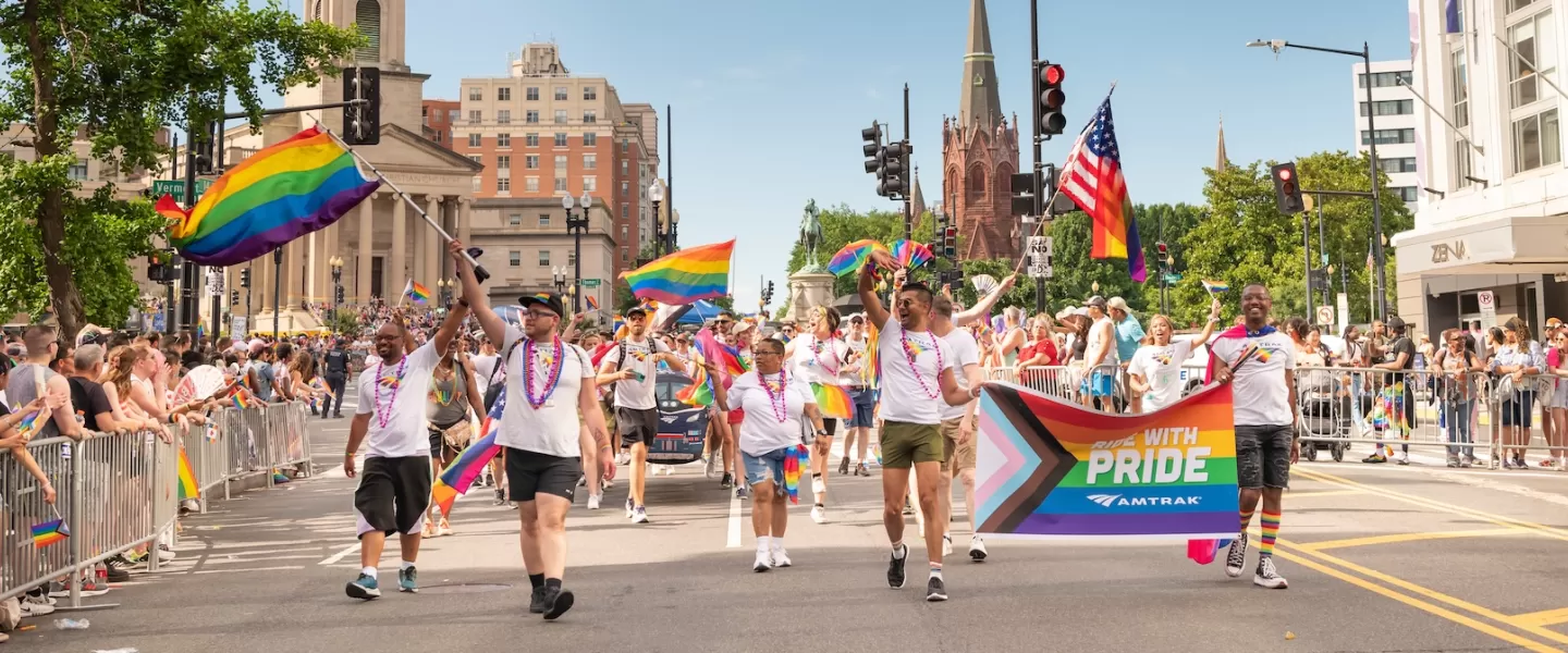 A vibrant group marches down a city street holding rainbow flags and a “Ride With Pride” banner during a Pride Parade, cheered on by festive crowds.