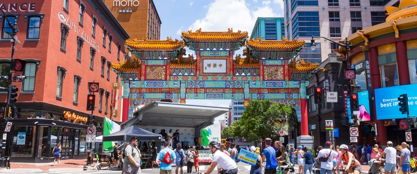 People walk and bike through a lively street festival beneath the colorful Friendship Archway in Washington, DC.