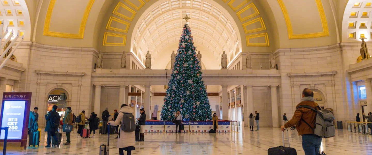 Visitors walk through the grand hall of Union Station in Washington, DC, where a towering decorated Christmas tree glows beneath the arched, gold-accented ceiling.