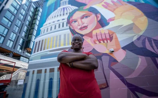 A person crosses their arms and smiles for a portrait in front of a colorful mural that depicts someone signing with the U.S. Capitol in the background. 

