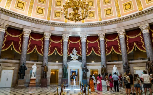 Visitors stand beneath the ornate dome and red curtains of the National Statuary Hall inside the U.S. Capitol.
