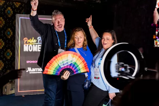 Three people enthusiastically pose with a rainbow fan at a Pride event photo booth, with a "Pride Proud Moments" backdrop behind them.